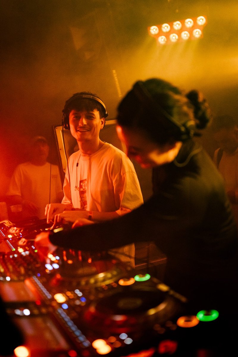 Robin smiling at Demi behind the decks, golden club lights